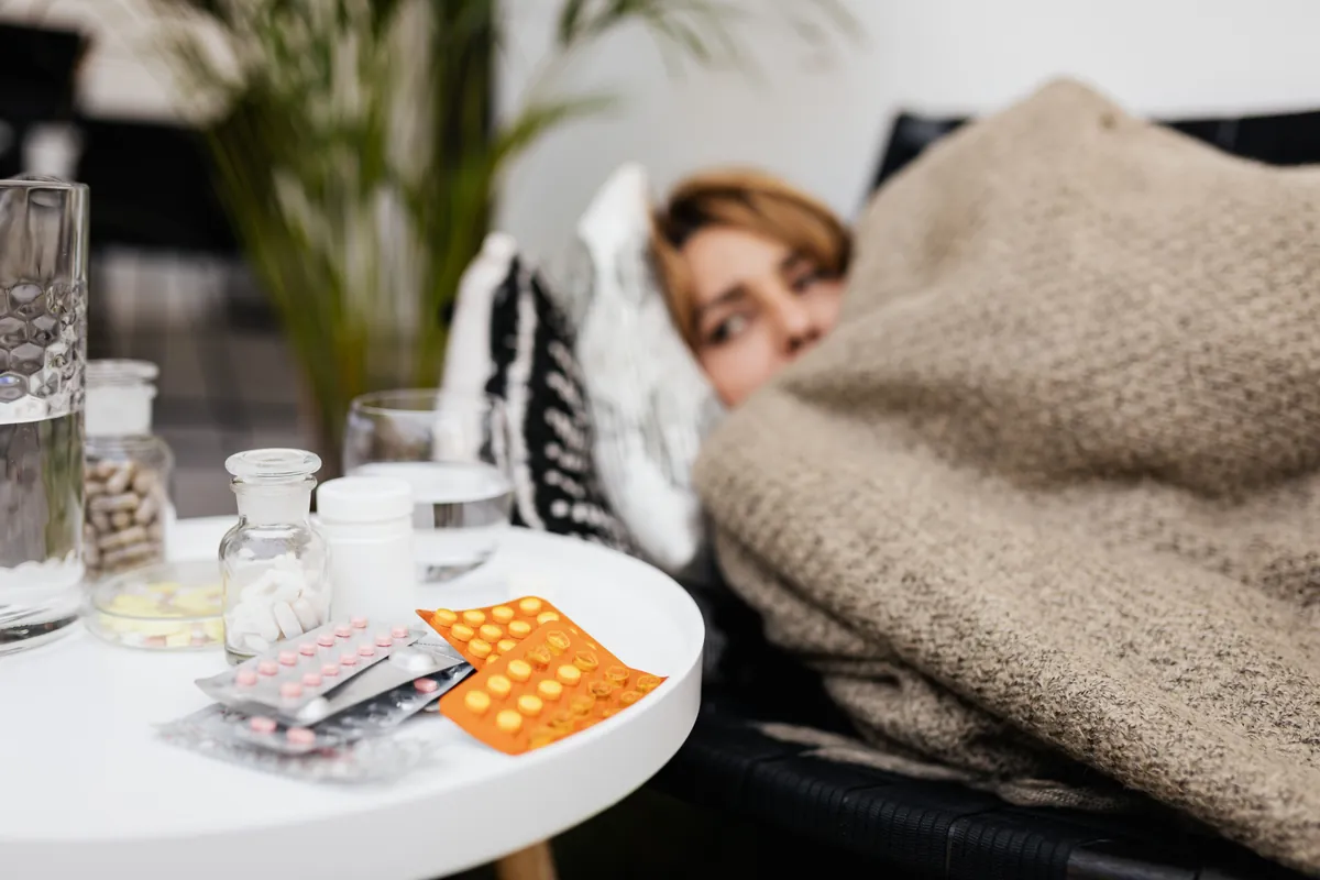 A woman lying on the couch under a blanket, next to a small table covered with pills, bottles and a glass of water.