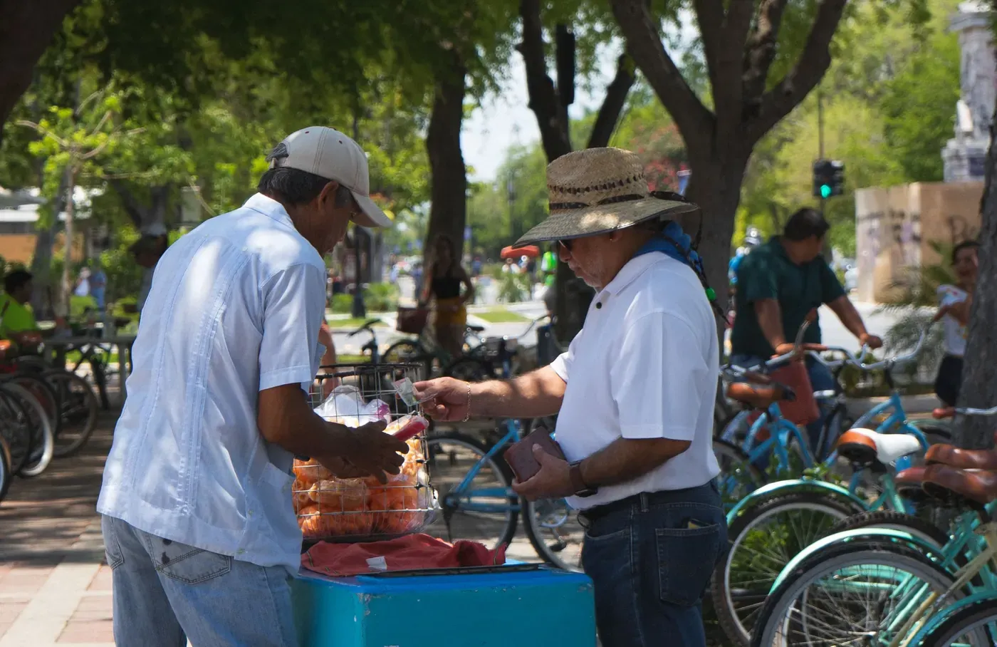 Una calle de Mérida a media tarde: un vendedor de paletas atiende a un señor de sombrero junto a su carrito, bajo los árboles y entre bicicletas estacionadas.