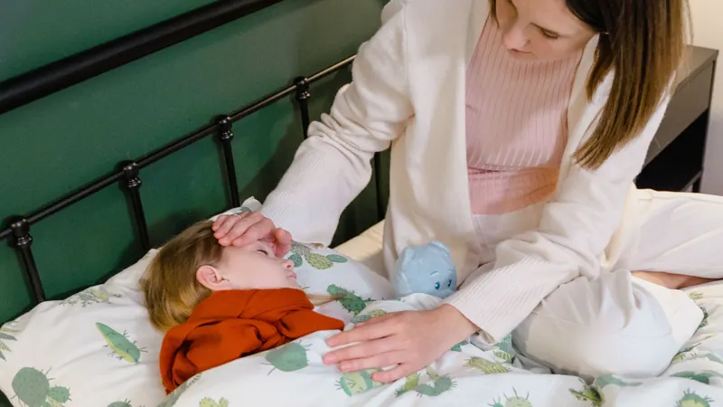 A mother sitting on the bed with her hand on the forehead of her son, who lies tucked in and looks unwell.