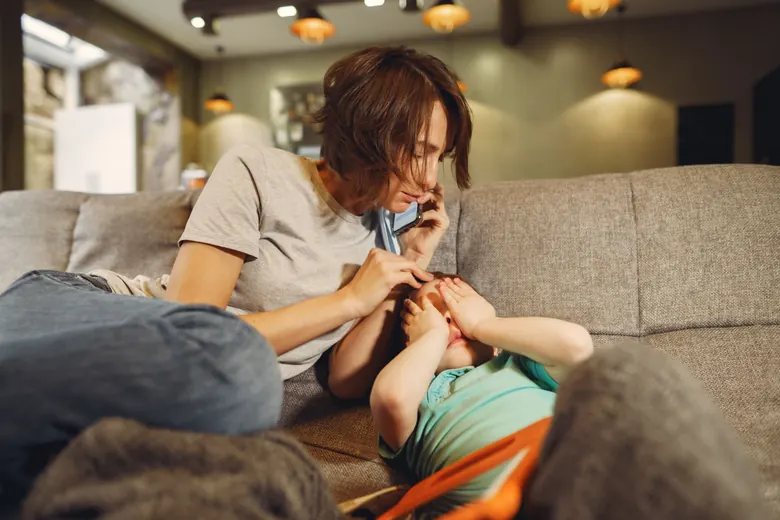 A mother talks on the phone sitting on her living-room couch, with her other hand resting on the head of her son, who lies beside her covering his eyes.